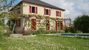 Edificio de piedra con contraventanas rojas y un jardín delantero.