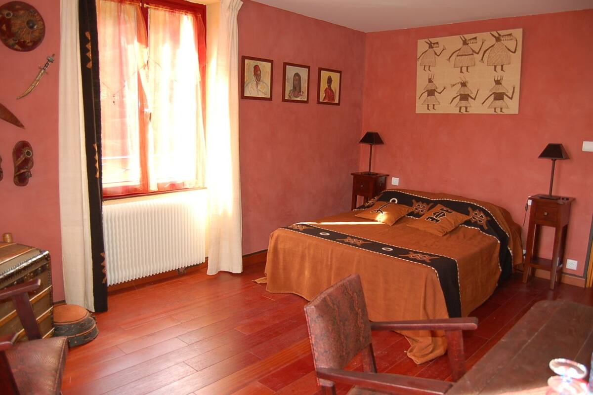 A bedroom with a bed, a chair, a table, a radiator, and a window with curtains.