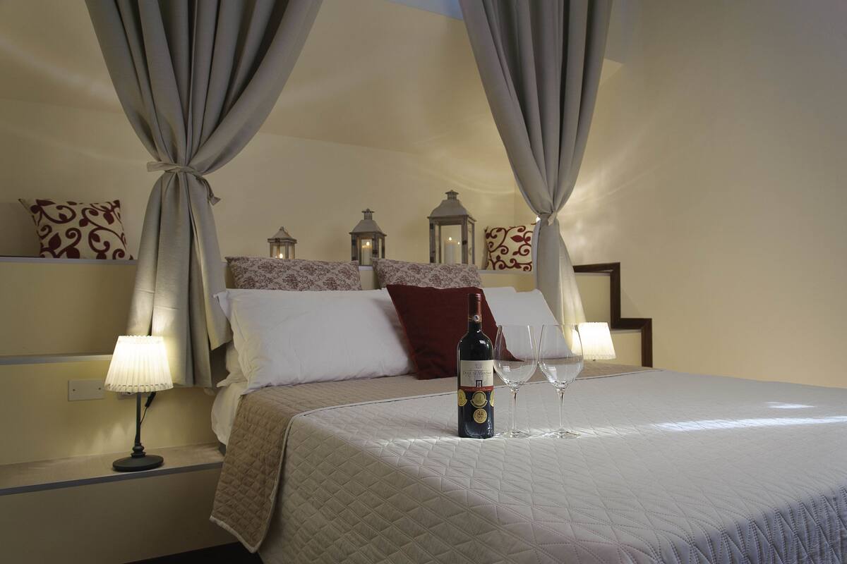 Un lit bien fait, avec une bouteille de vin et deux verres, une lampe sur la table de chevet et des lanternes décoratives sur la tête de lit.