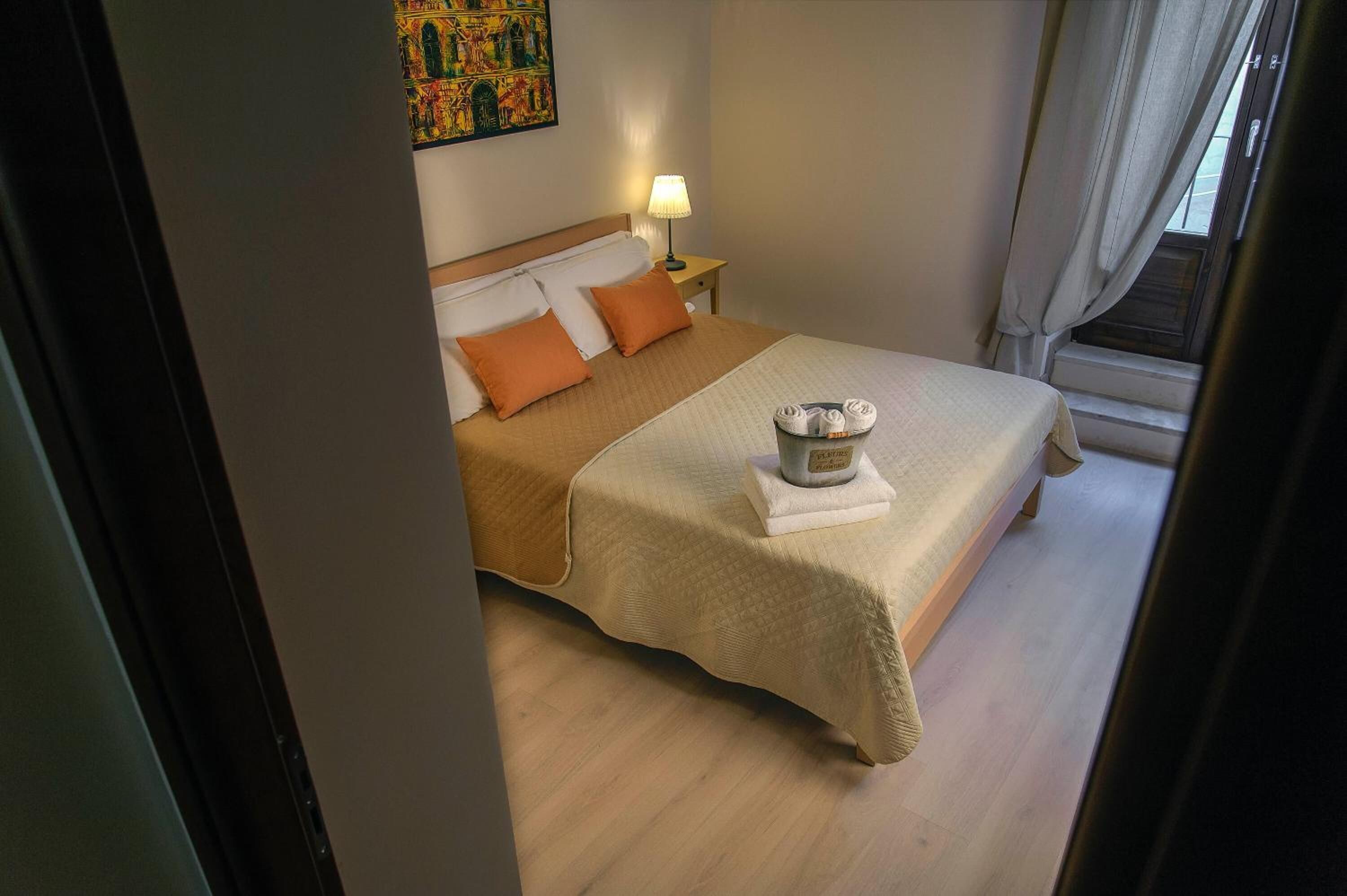 A neatly made bed with a beige bedspread and two orange pillows, a bedside lamp, and a decorative item on the bed.