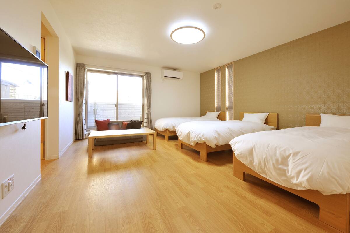 A hotel room with three single beds, a wooden floor, a large window with curtains, and a view of the outside.
