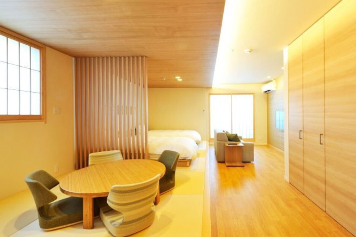 A modern hotel room with a wooden floor, a round dining table with chairs, a bed, a sofa, and a wardrobe.