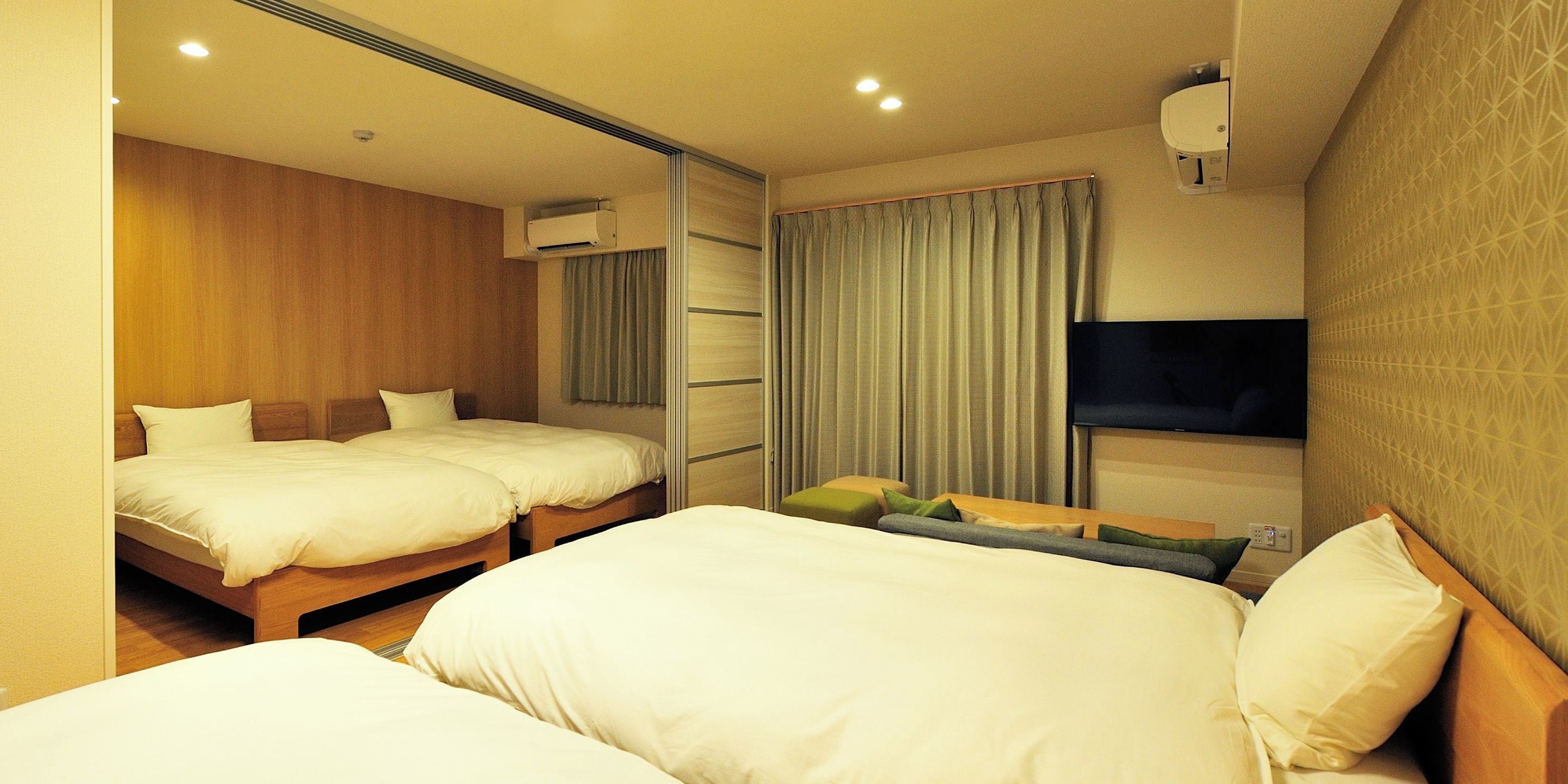 A hotel room with two beds, a flat-screen TV, an air conditioning unit, and a window with curtains.