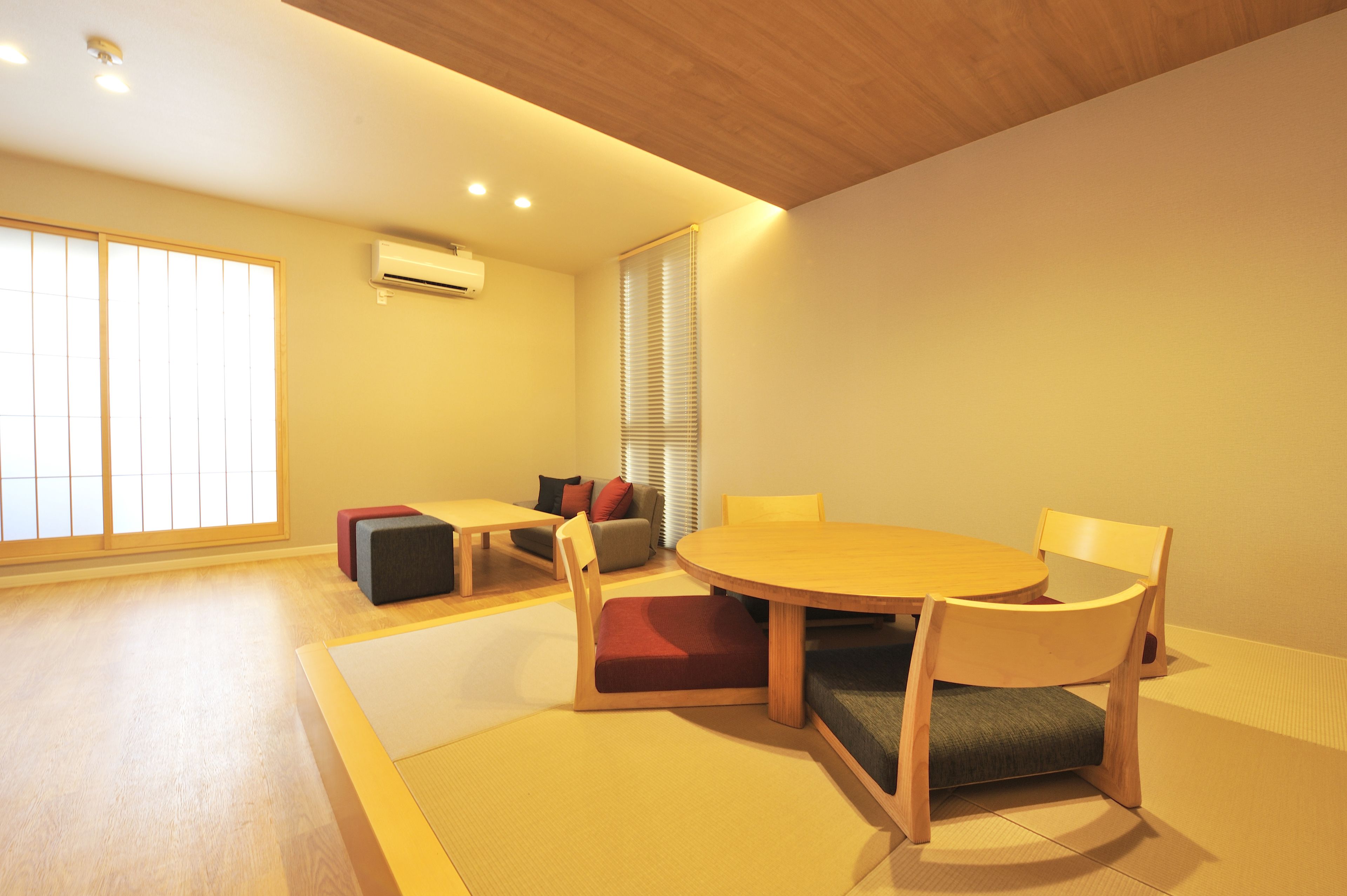Una habitación con esteras de tatami, una mesa redonda de madera y sillas. Hay un sof á con cojines y una ventana con persianas.