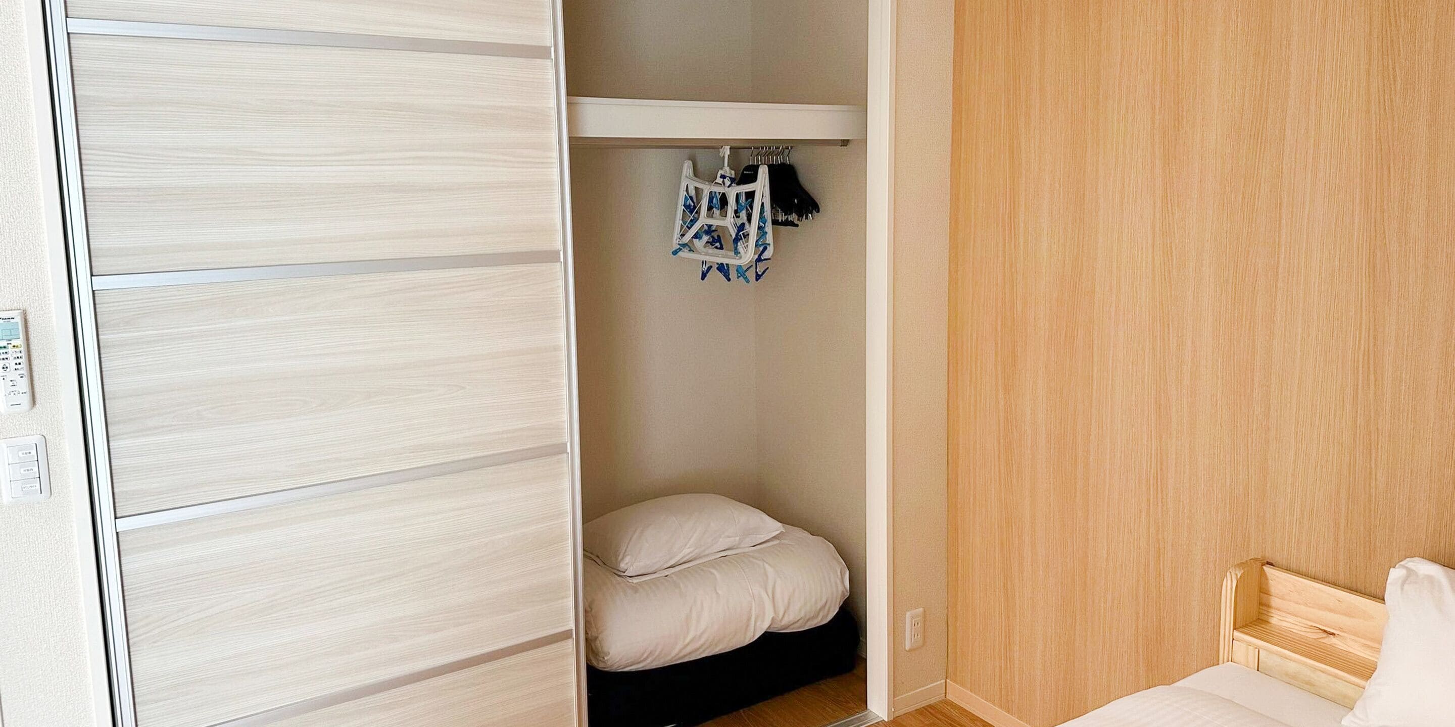 Een slaapkamer met een houten garderobe, een bed met wit beddengoed en een klein plankje met een tas met blauw-wit patroon.