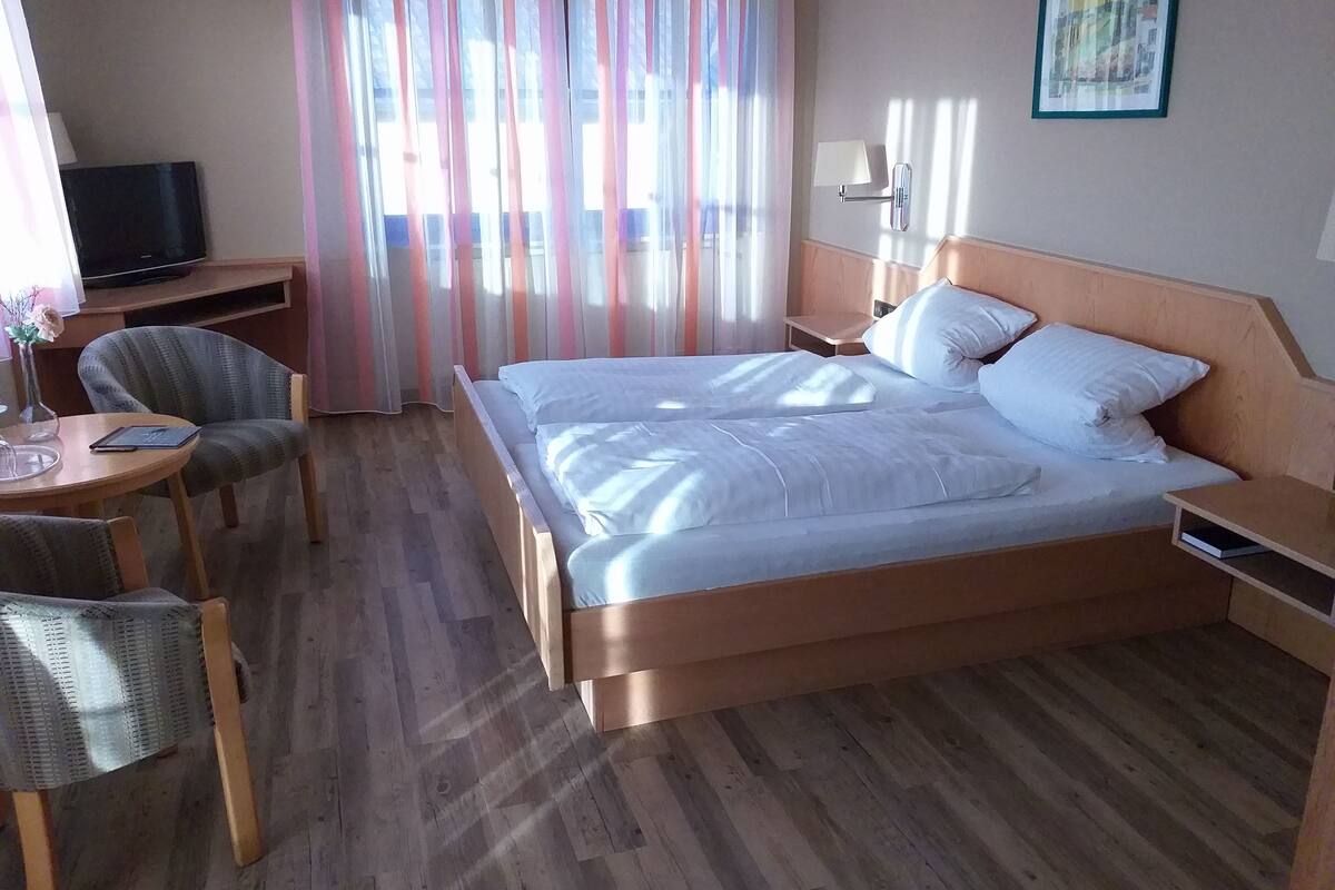 Ein Hotelzimmer mit einem hölzernen Bett, einem Schreibtisch mit Stuhl, einem Fernseher und einem Fenster mit leichtem Vorhang.