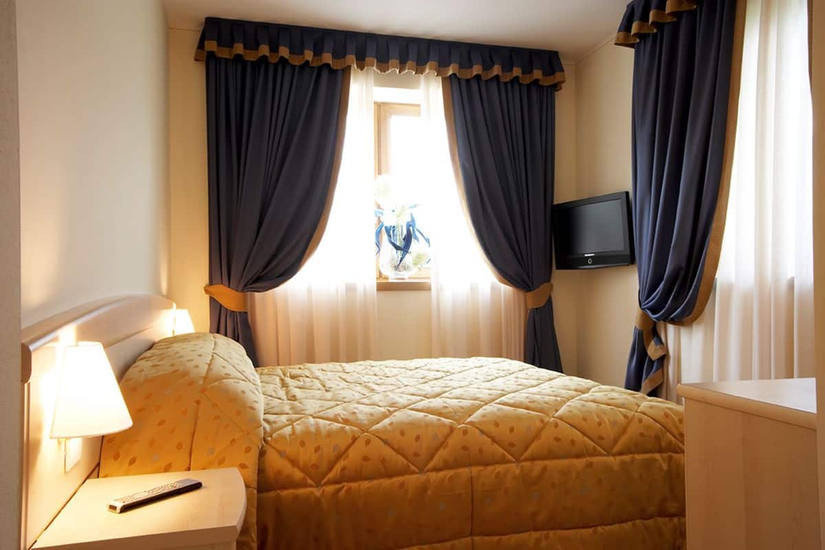 Una habitación de hotel con cama, mesita de noche, control remoto, televisor en la pared y cortinas.
