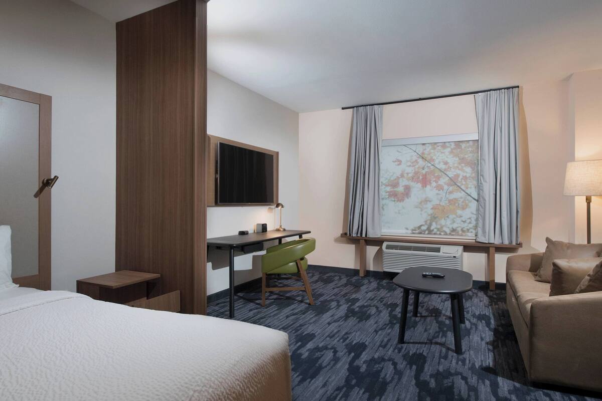 A hotel room with a bed, a desk, a chair, a small table, a sofa, a TV, and a window with curtains.