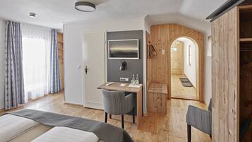 Ein modernes Hotelzimmer mit Bett, Schreibtisch und Stuhl. An der Wand ist ein Fernseher befestigt, es gibt einen Holzschrank und eine Holztür zu einem weiteren Raum.