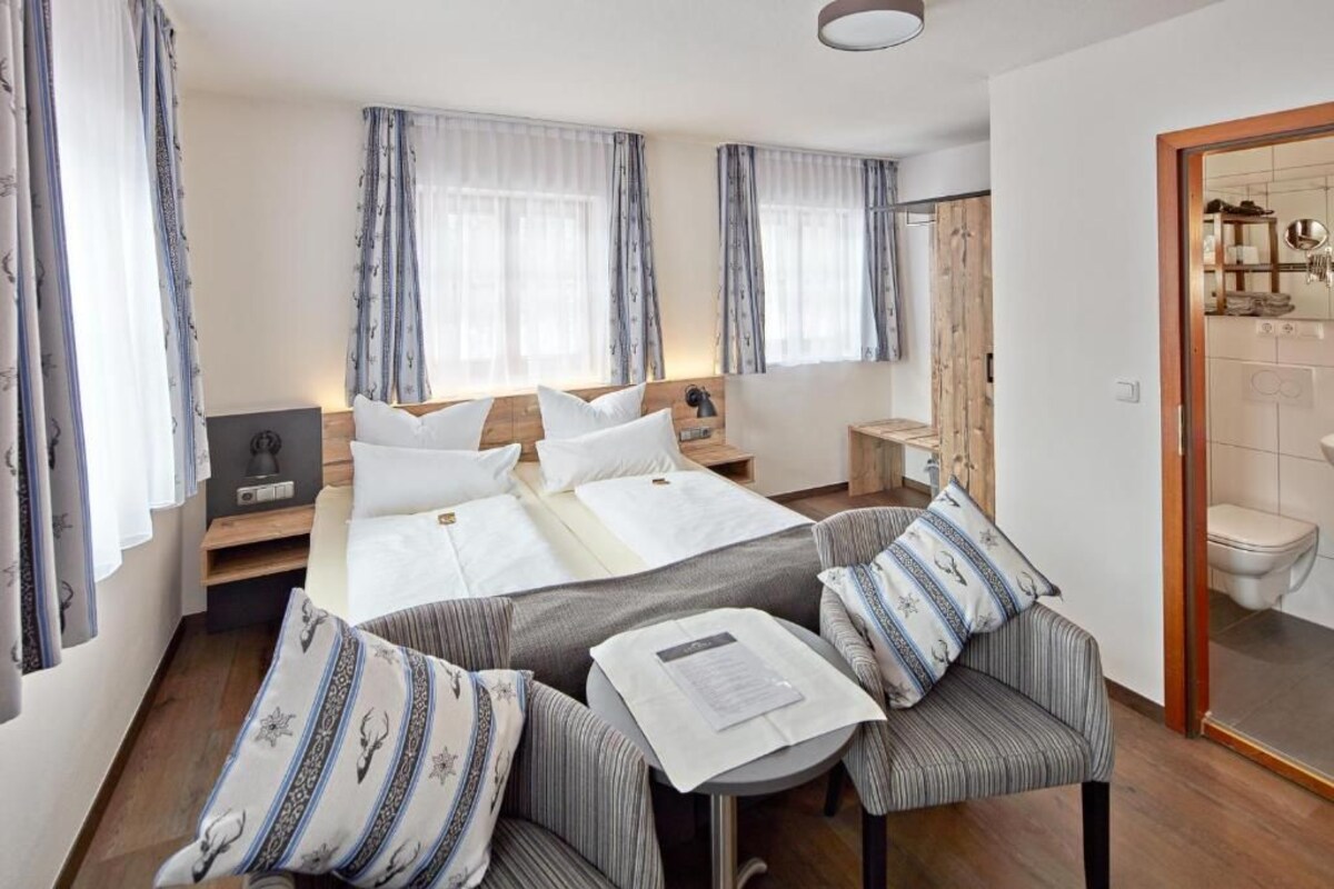 A hotel room with a bed, two bedside tables, a small table with a chair, and a bathroom visible through an open door.