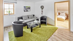 A modern living room with a sofa, a coffee table, and a chair. There is a window with blinds, a painting on the wall, and a plant on the windowsill. - Alte Säge Ruhpolding (Ruhpolding)