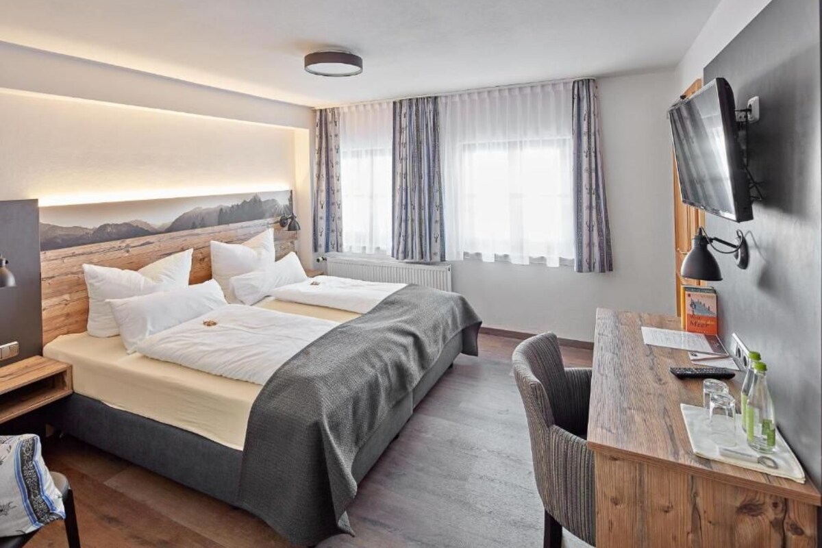 Quarto de hotel com cama, escrivaninha, cadeira, televisão e janela com cortinas.