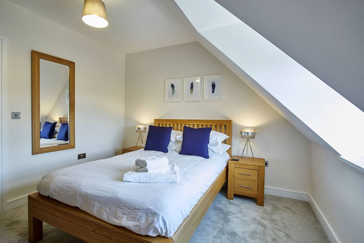 A bedroom with a wooden bed, white bedding, two blue pillows, a nightstand, and a mirror.