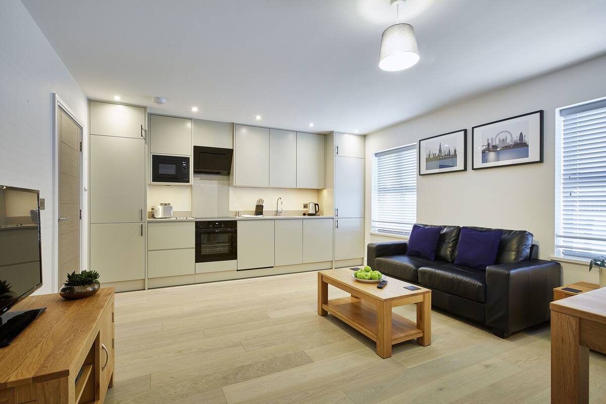 A modern living room with a sofa, coffee table, and a kitchen area with cabinets and appliances.