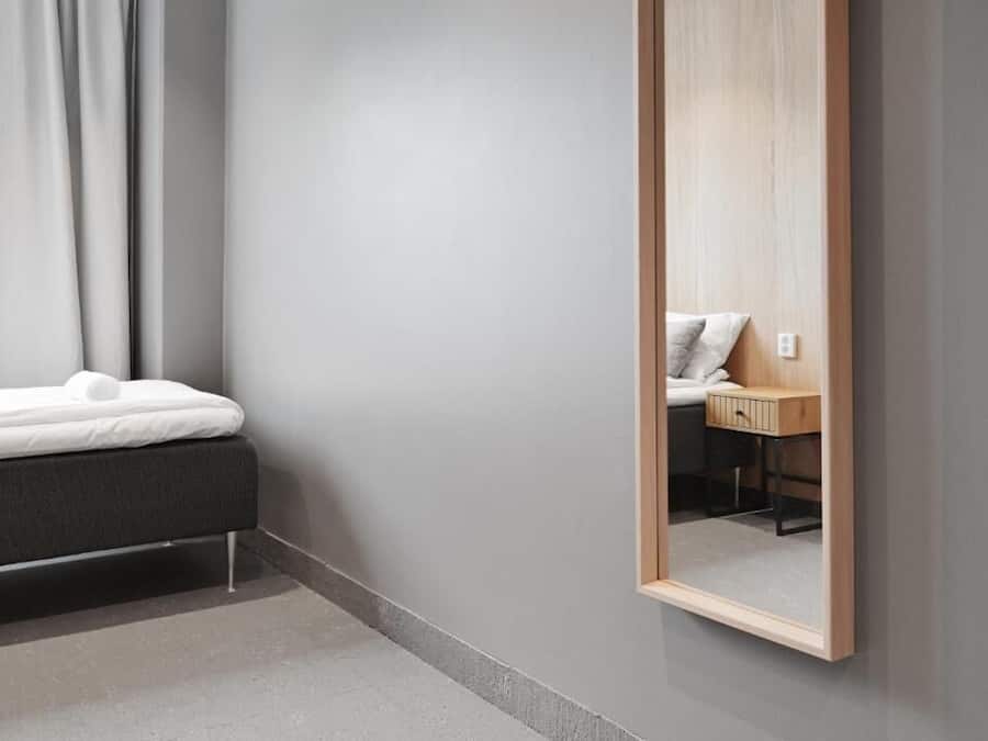 A modern bedroom with a bed, a bedside table, and a mirror reflecting a bathroom with a sink and cabinet.