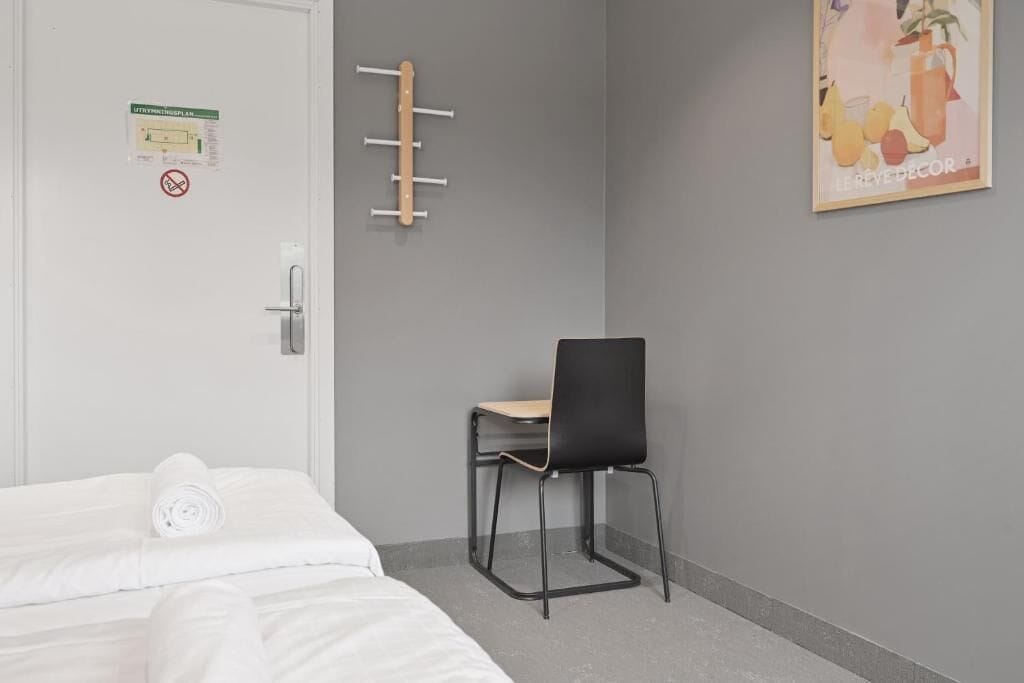 Ett hotellrum med en säng, en stol, en klädställning och en affisch på väggen.