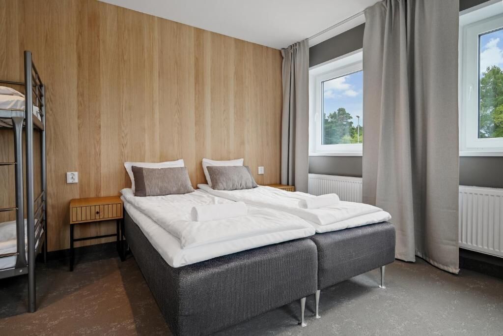 Ein modernes Schlafzimmer mit Etagenbett, einem großen Bett mit grauem gepolsterten Kopfteil, einer Holzwand, einem Fenster mit Vorhängen und einer Heizung.