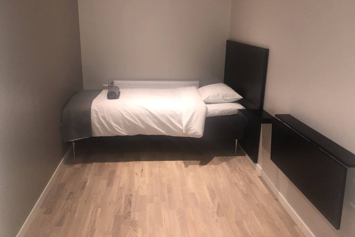 Una habitación pequeña con una cama individual, piso de madera, colcha blanca y cabecera negra.