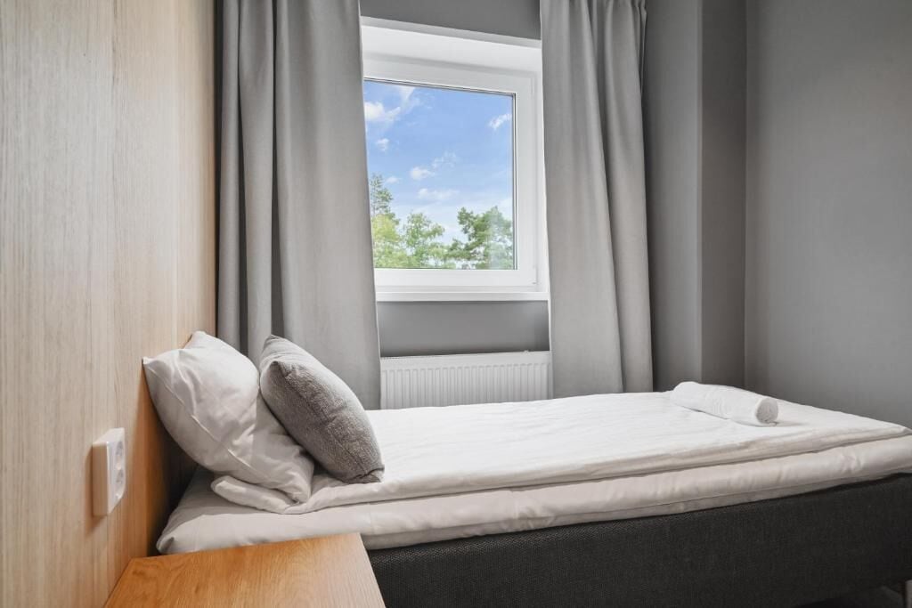 Una cama individual con colchón blanco, cortinas grises y ventana con vista a árboles.
