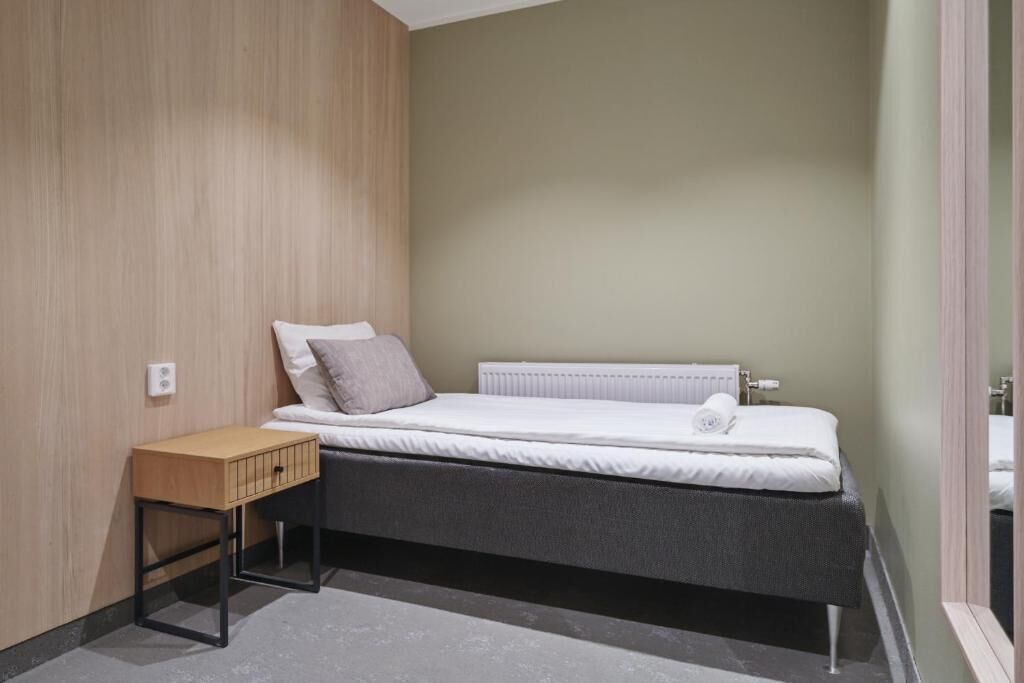 A small, single-bed room with a wooden panel wall, a bedside table, and a radiator.