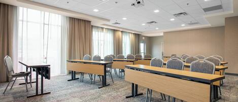A conference room with multiple tables and chairs, large windows with curtains, and a carpeted floor.