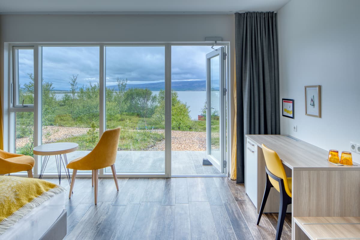 A modern room with a large sliding glass door leading to a balcony with a view of a lake and trees. There is a bed, a desk, and two chairs.