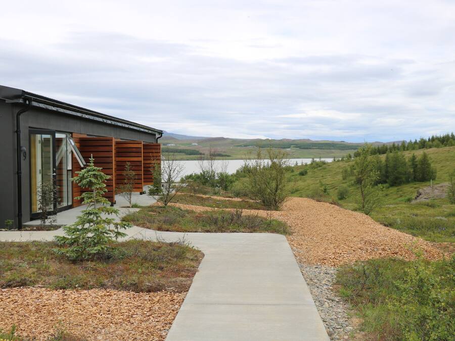 A modern house with a concrete pathway leading to a wooden deck, surrounded by a natural landscape with a lake and hills in the background.