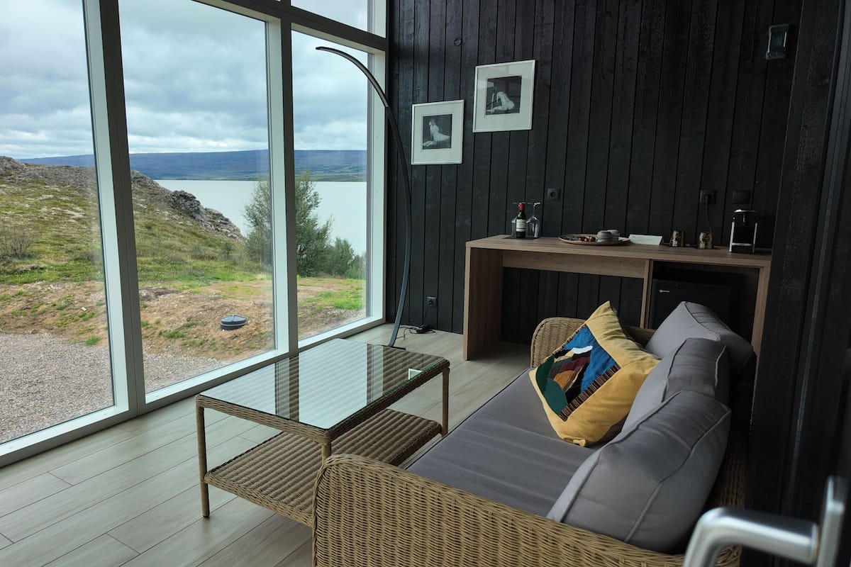A modern room with large windows offering a view of a lake and hills. It includes a sofa, a small table, and a wicker chair.