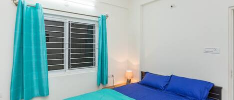 A bedroom with a bed, a window with curtains, a nightstand with a lamp, and a ceiling fan.