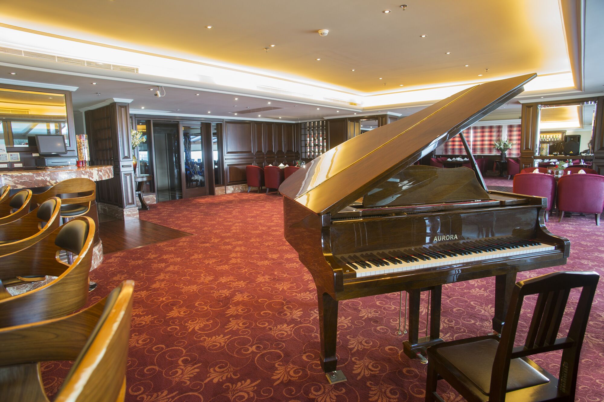 Un piano de cua majestuós en una habitació àmplia, amb una catifa vermella, cadires de fusta i una zona de bar al fons.
