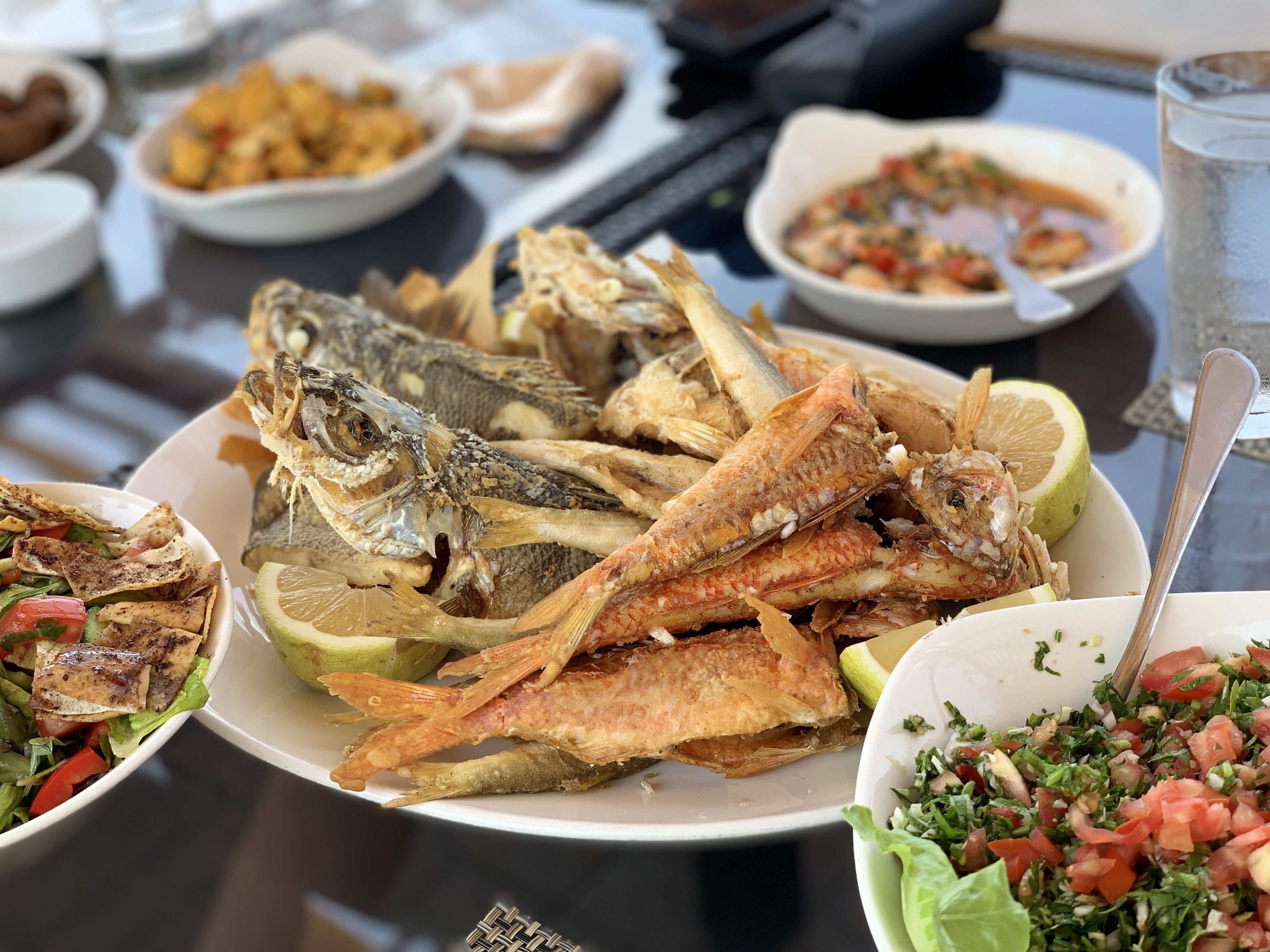 A table with various dishes including fried fish, salads, and a bowl of chopped vegetables.