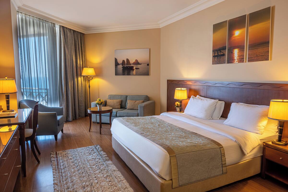 A hotel room with a large bed, a desk, a sofa, and a view of the sea through the window.