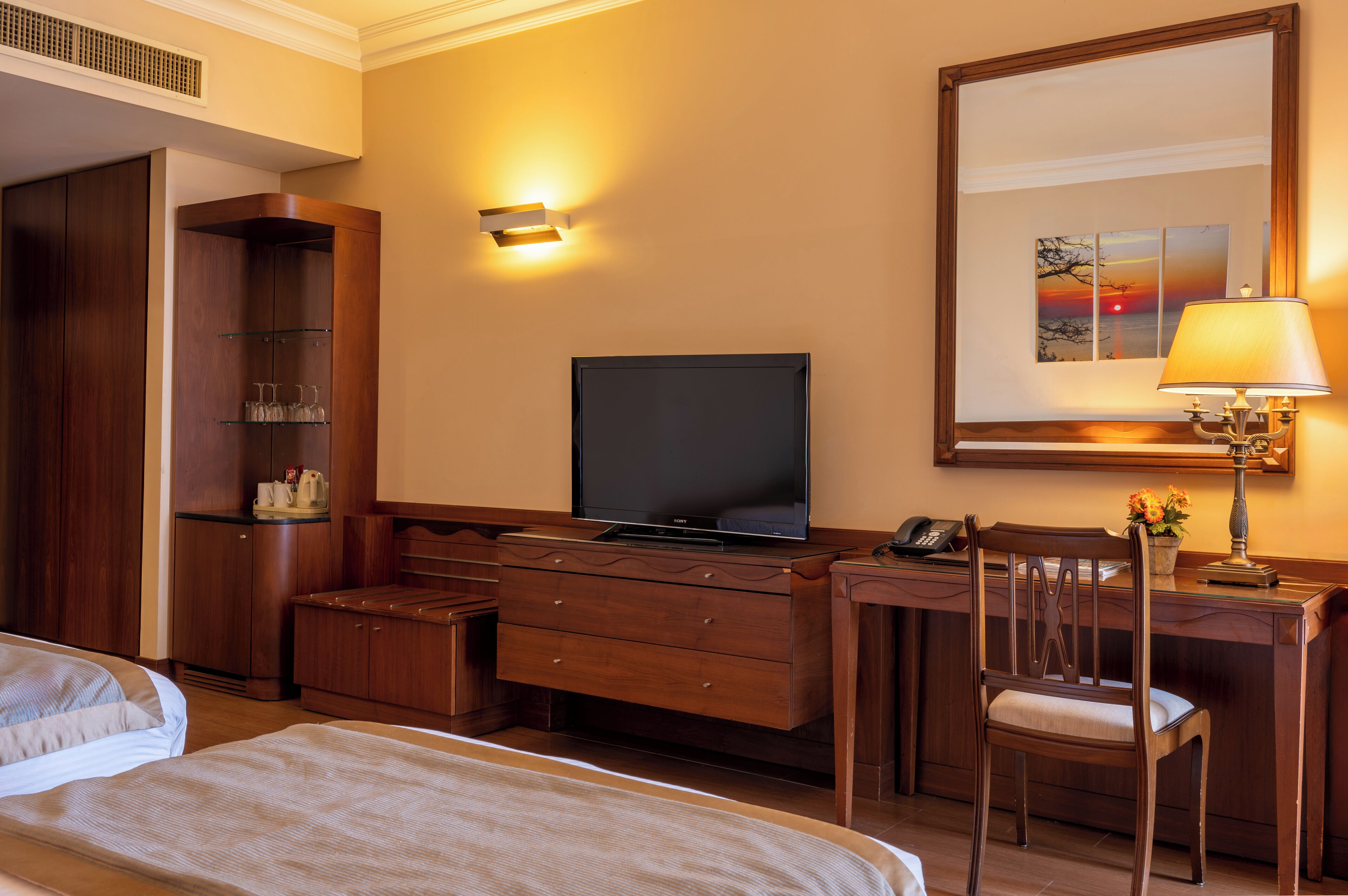 Habitación de hotel con una cama, un escritorio con televisor, una silla, una lámpara y un estante en la pared con vasos y una cafetera.