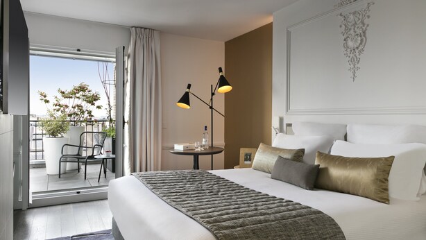 Une chambre d’hôtel avec un grand lit, une vue sur l’extérieur, une petite table avec une lampe et un balcon avec des chaises et un pot de fleurs.