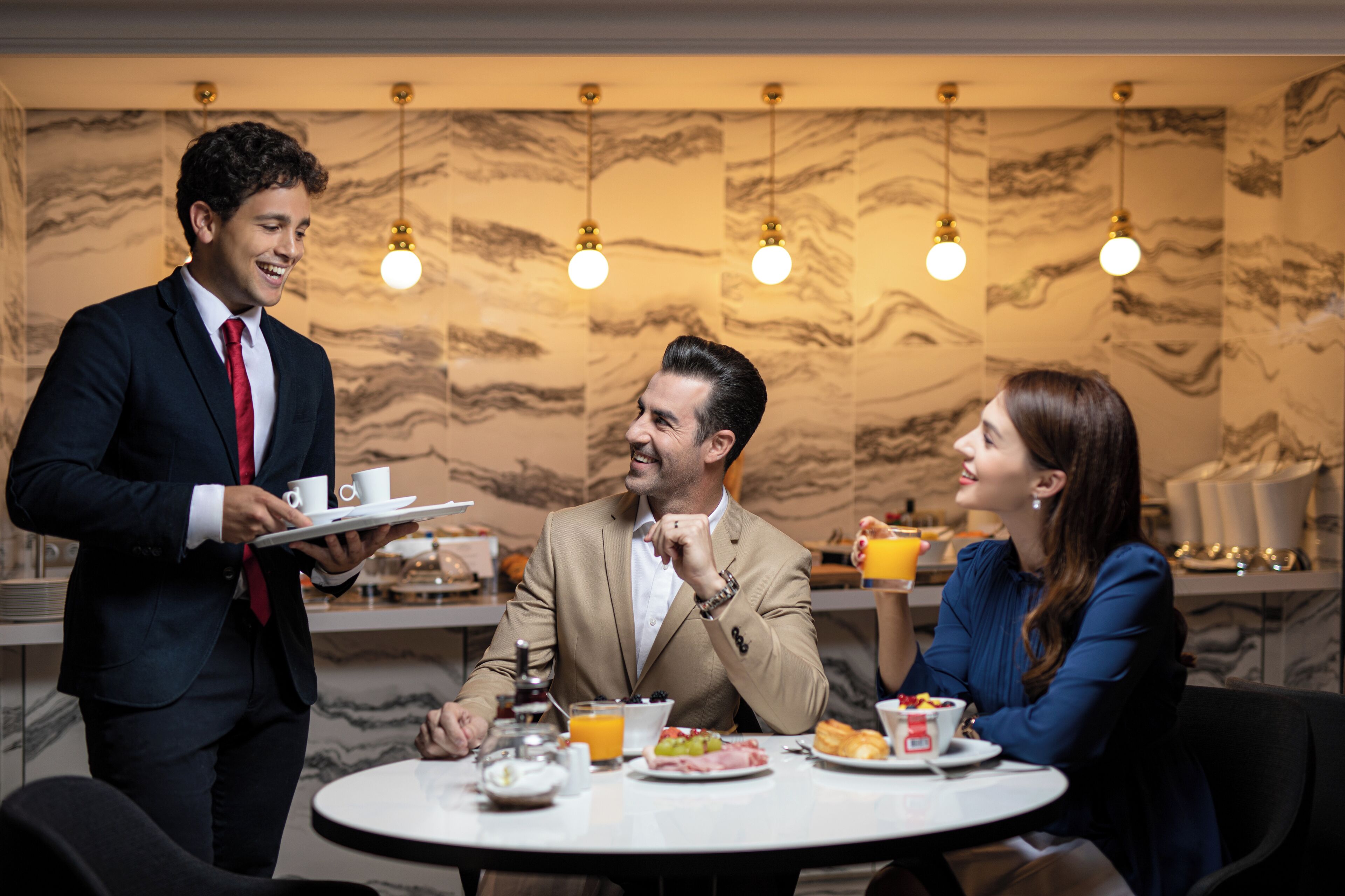 Comedor de hotel con pared de mármol, iluminación colgante y una mesa redonda con alimentos para el desayuno.