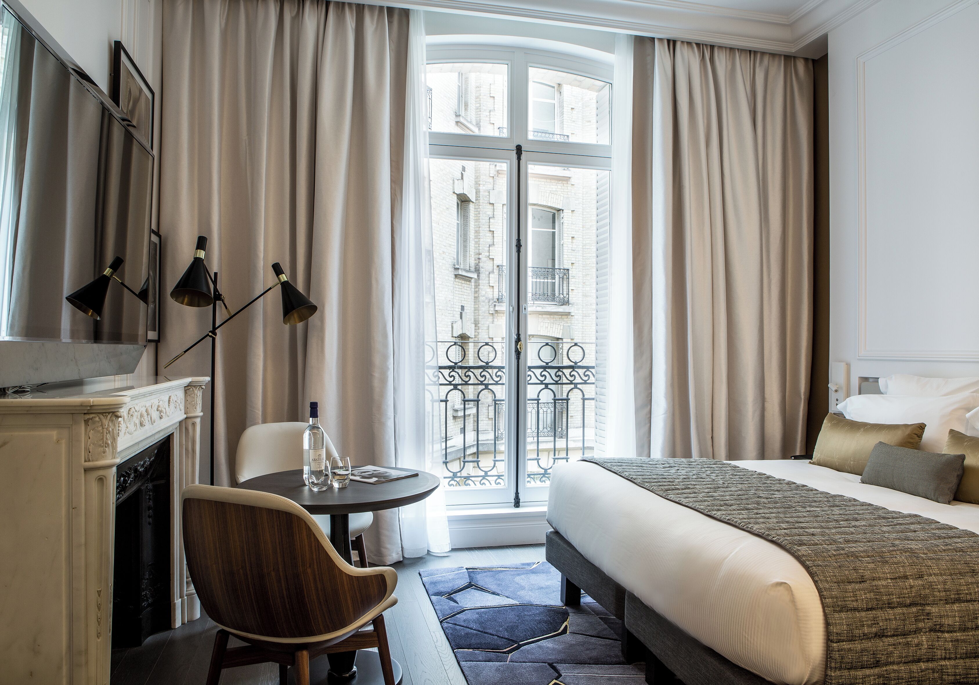 A hotel room with a bed, a desk with a chair, a fireplace, and a view of the city through a large window.