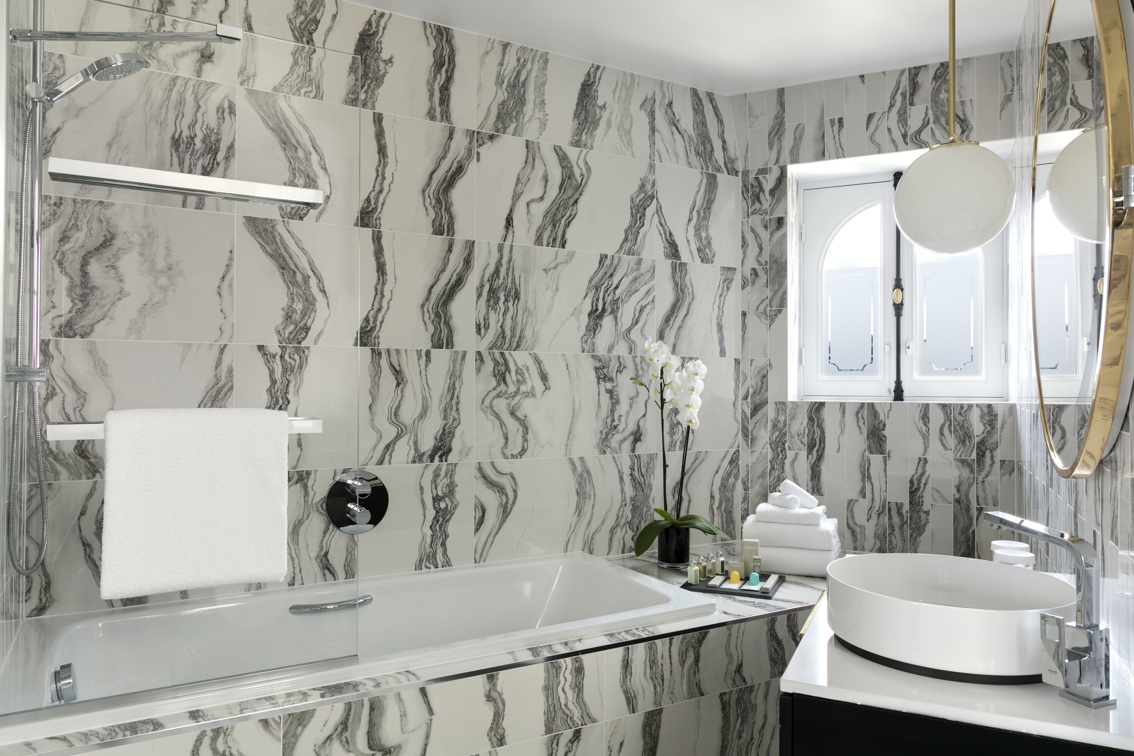 Un baño moderno con una bañera acristalada, un lavamanos blanco y paredes con diseño de mármol.