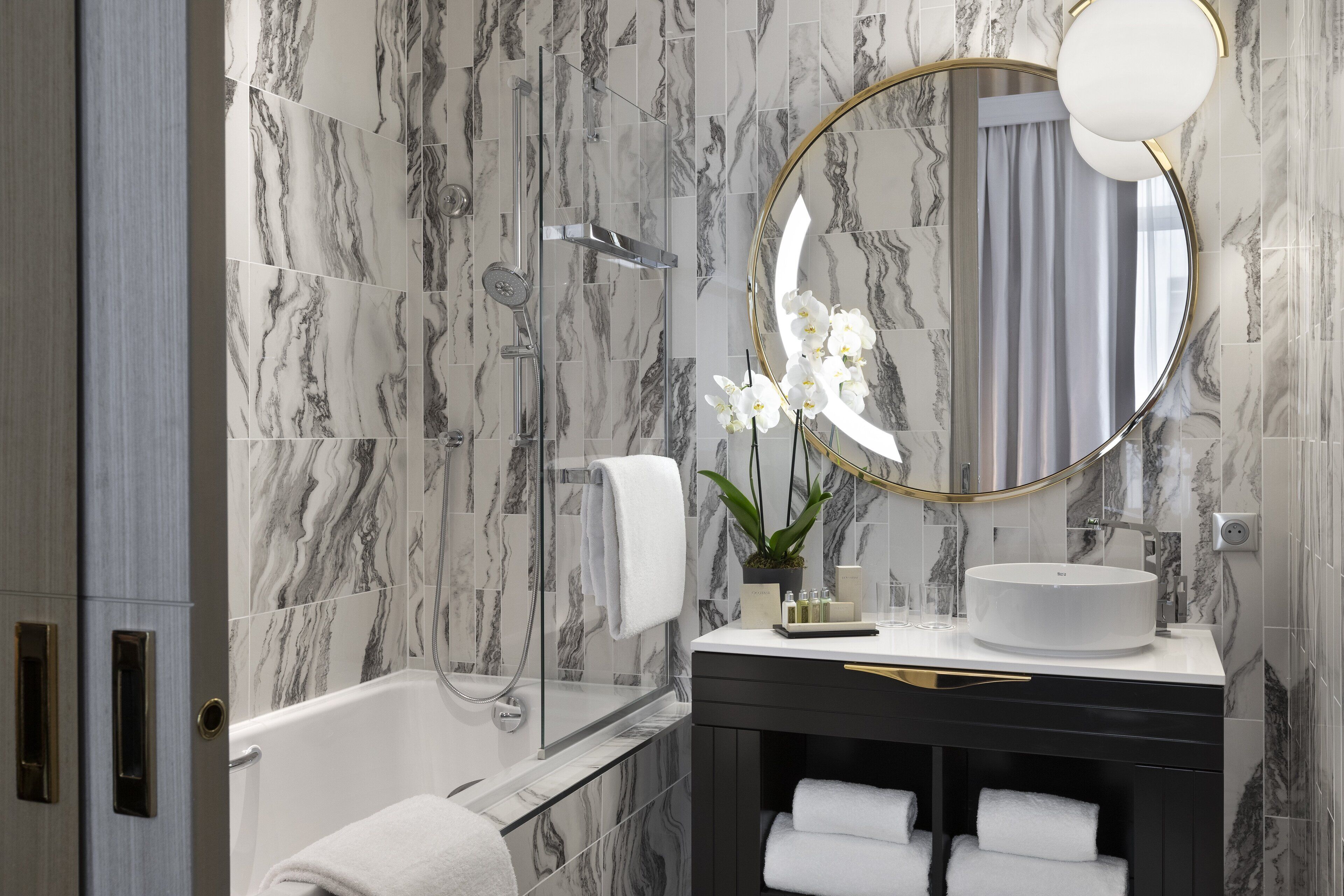 A modern bathroom with a marble backsplash, a round mirror, a white sink, and a bathtub.