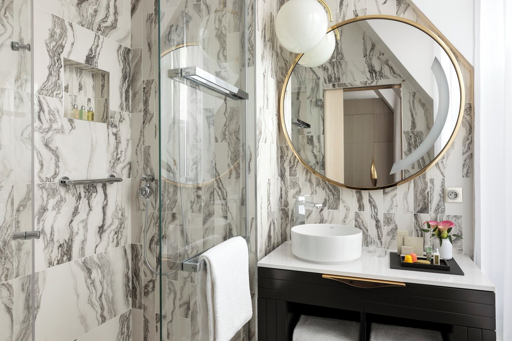 A bathroom with a glass-enclosed shower, a white sink, a round mirror with a gold frame, and marble-patterned walls.