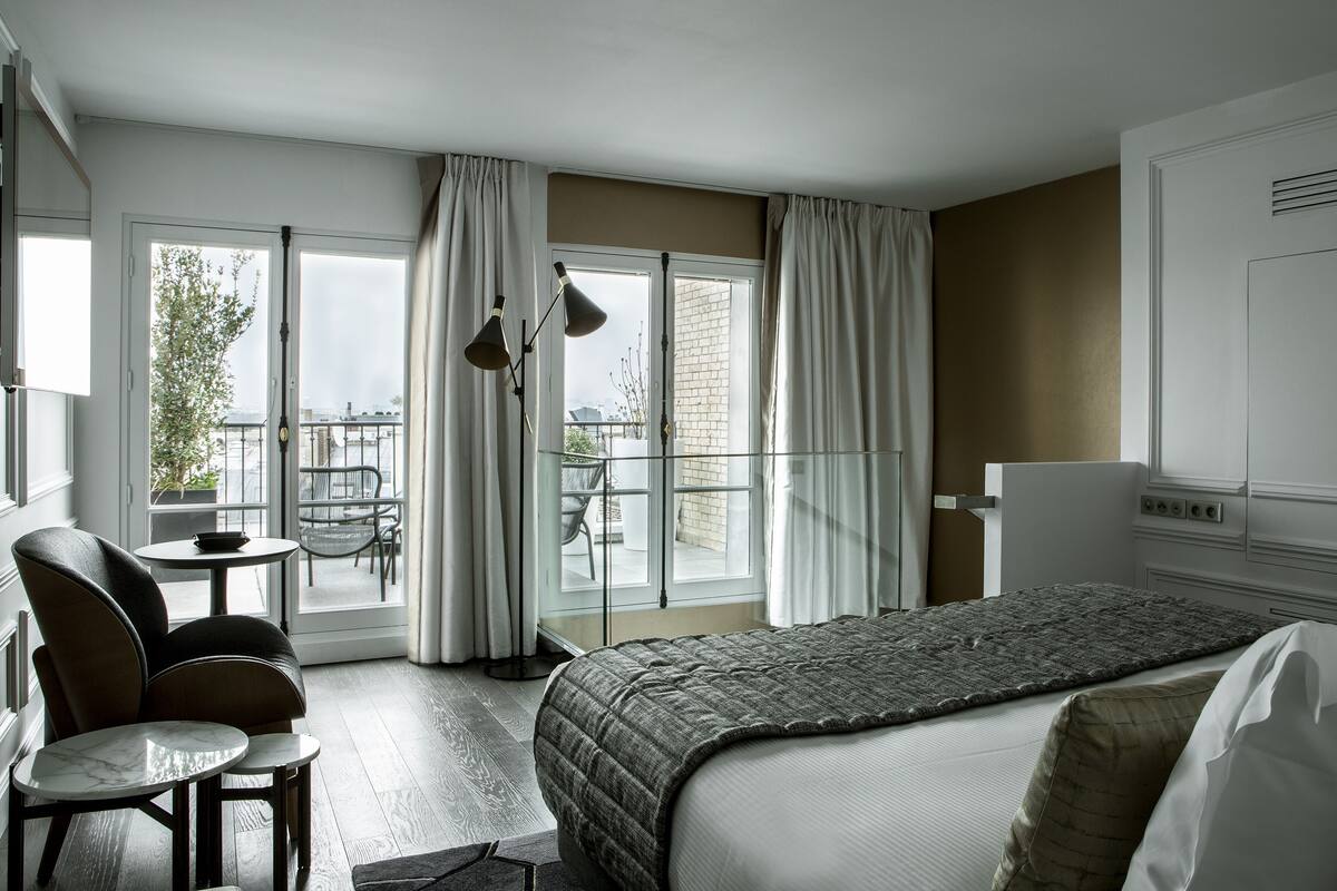 Hotelkamer met een bed, een stoel, een klein tafeltje en een balkon met gordijnen.