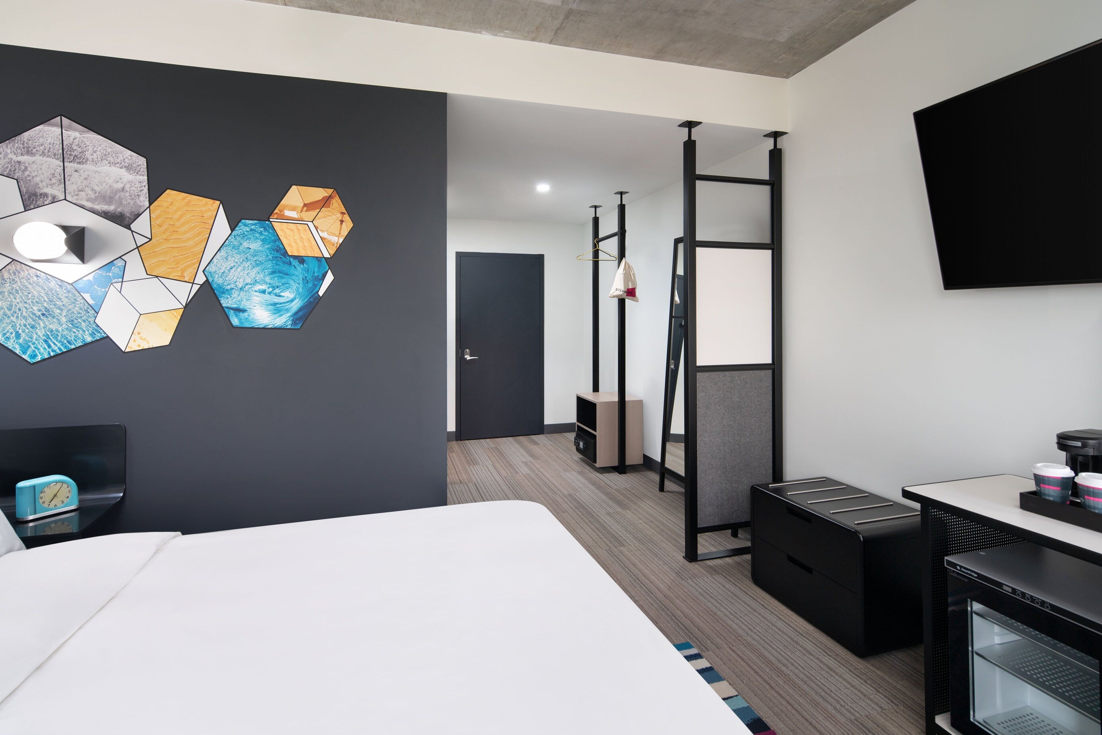 A modern hotel room with a large bed, a desk with a computer, a TV mounted on the wall, and a unique hexagonal wall art piece.