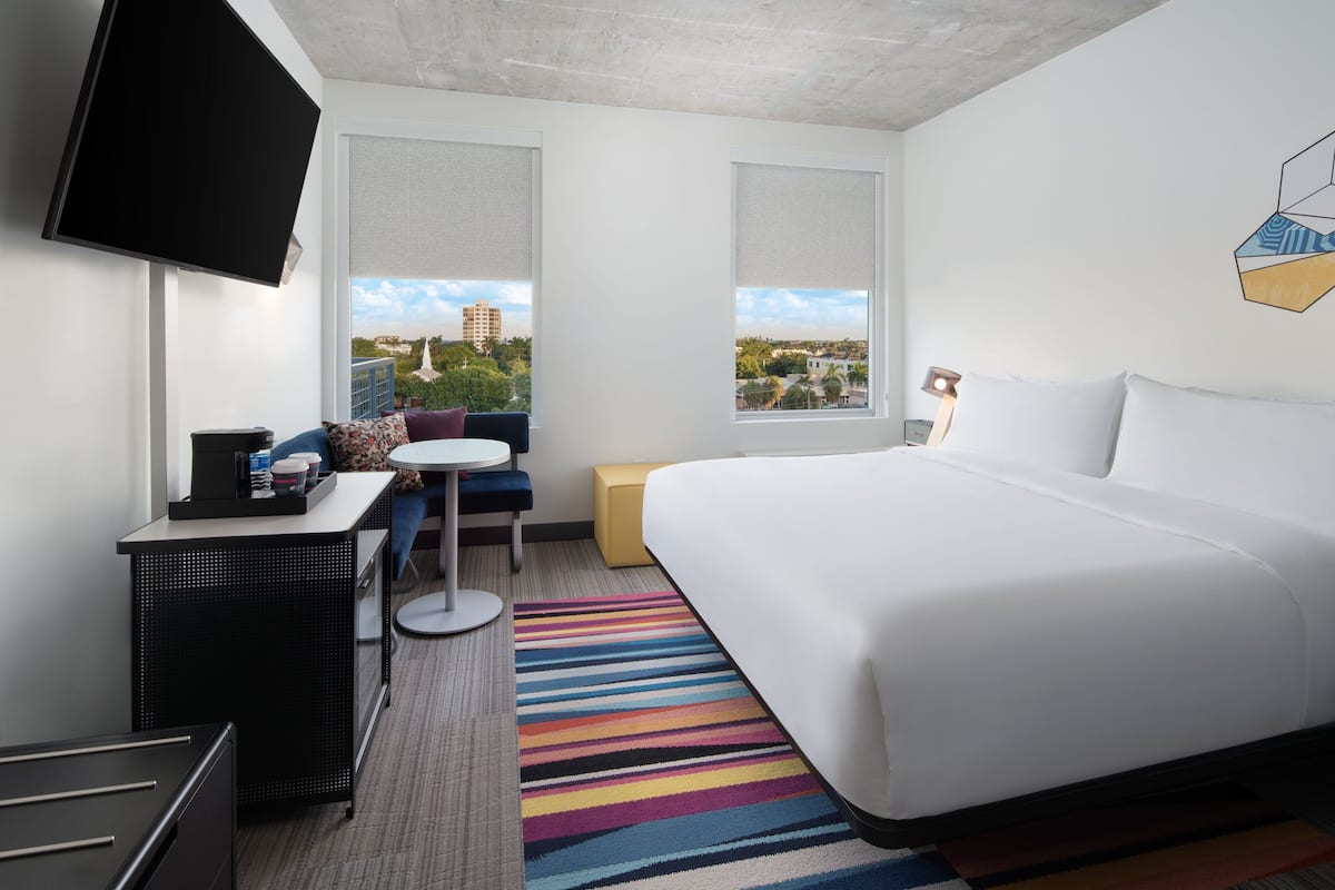 A hotel room with a large bed, a desk with a chair, a TV mounted on the wall, and a window with a view of buildings and trees.