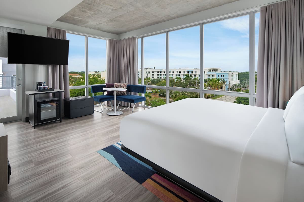 A modern hotel room with a large bed, a flat-screen TV mounted on the wall, a small desk with a coffee maker, and a balcony with a view of the city.