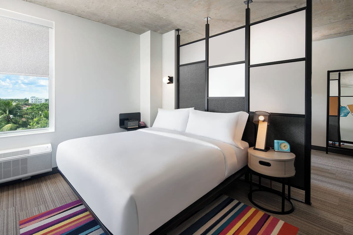 A modern hotel room with a large bed, a bedside table with a lamp, a small clock, and a view of greenery outside.