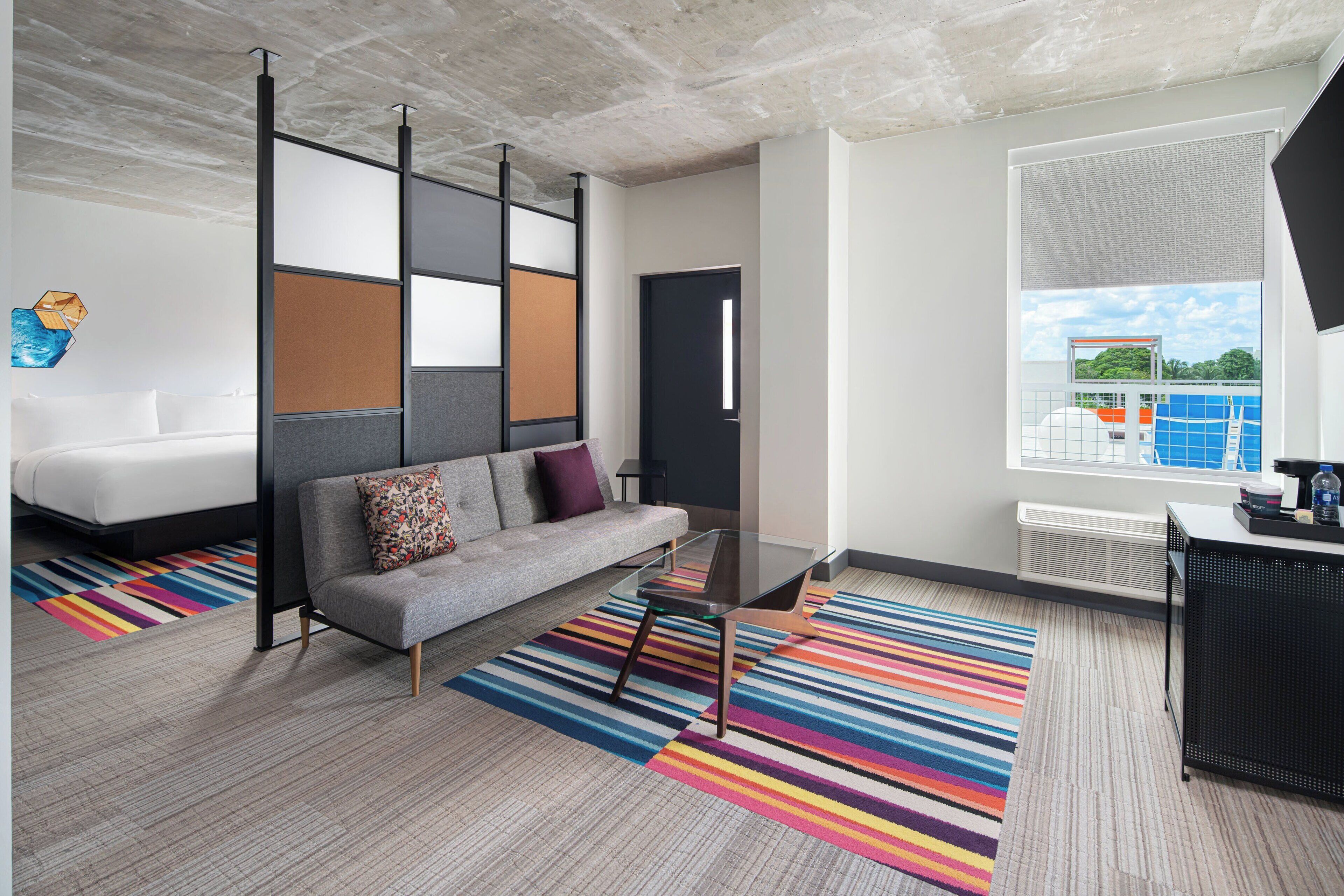 Een moderne hotelkamer met een grijze bank, een glazen salontafel, een kleurrijk gestreept vloerkleed en een groot raam met uitzicht op een blauw zwembad en groen.
