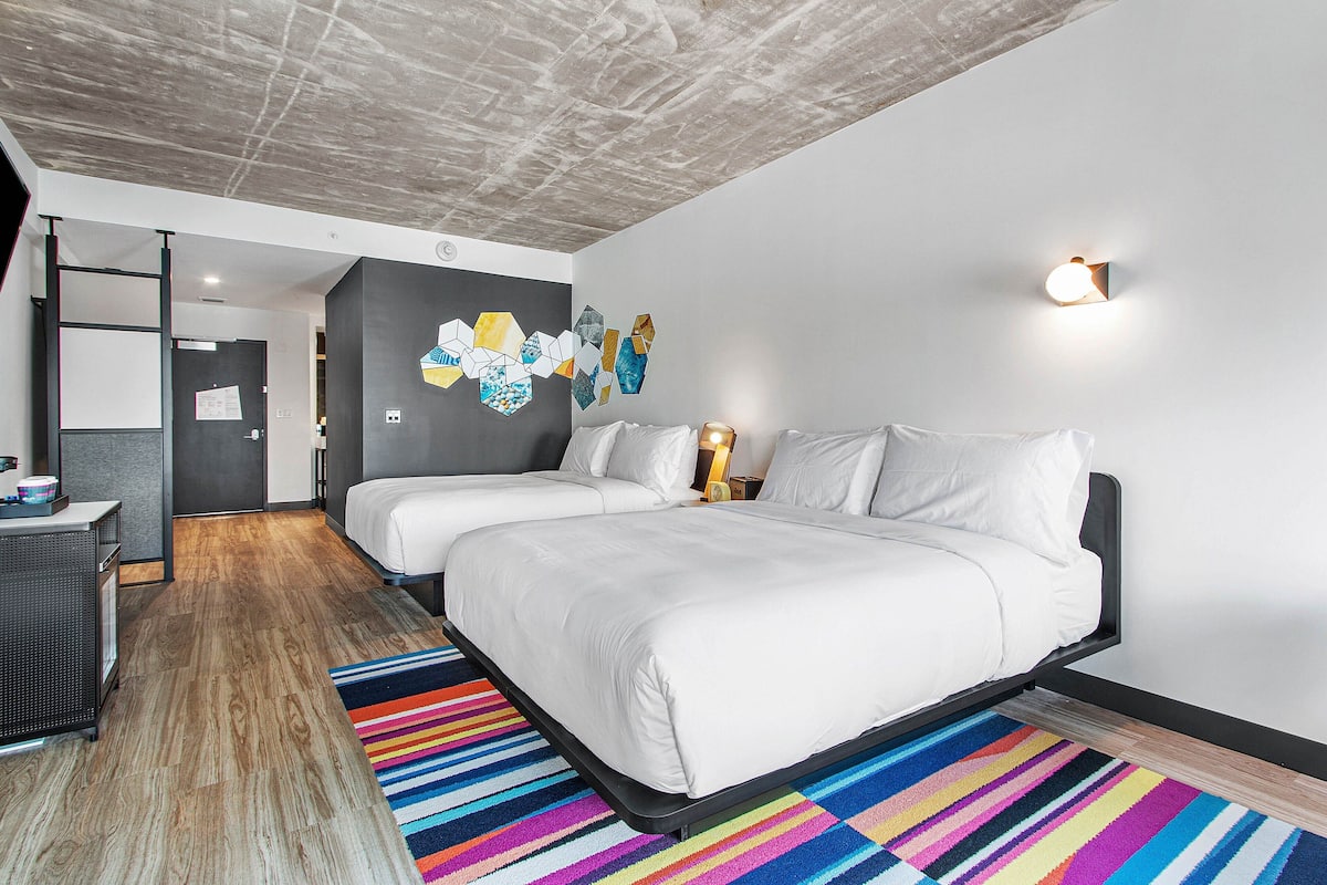 A hotel room with a bed, two bedside tables, a desk, a colorful striped rug, and a geometric wall art piece.
