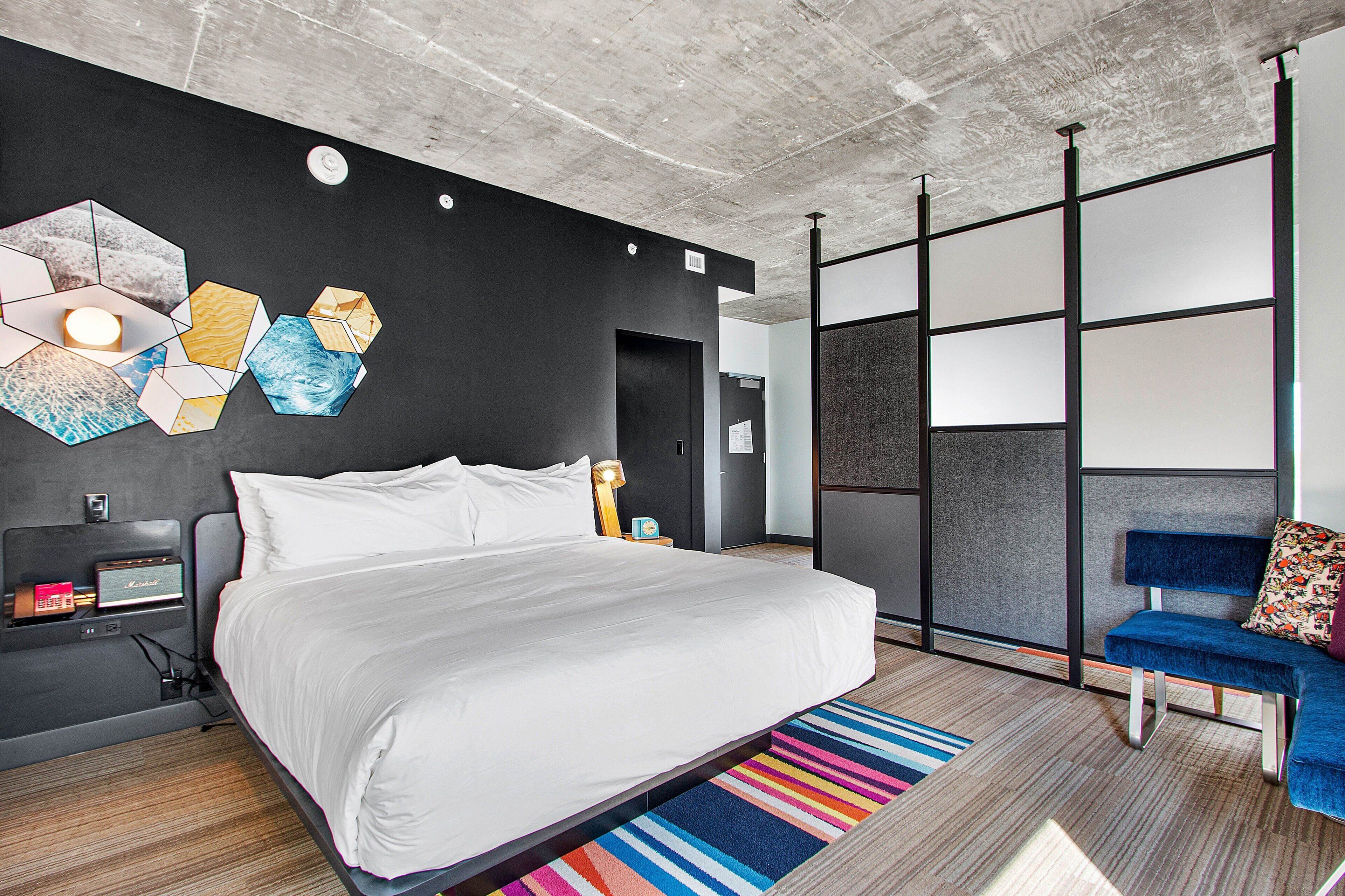 A modern hotel room with a large bed, a desk with a lamp, a blue chair, and a colorful rug.