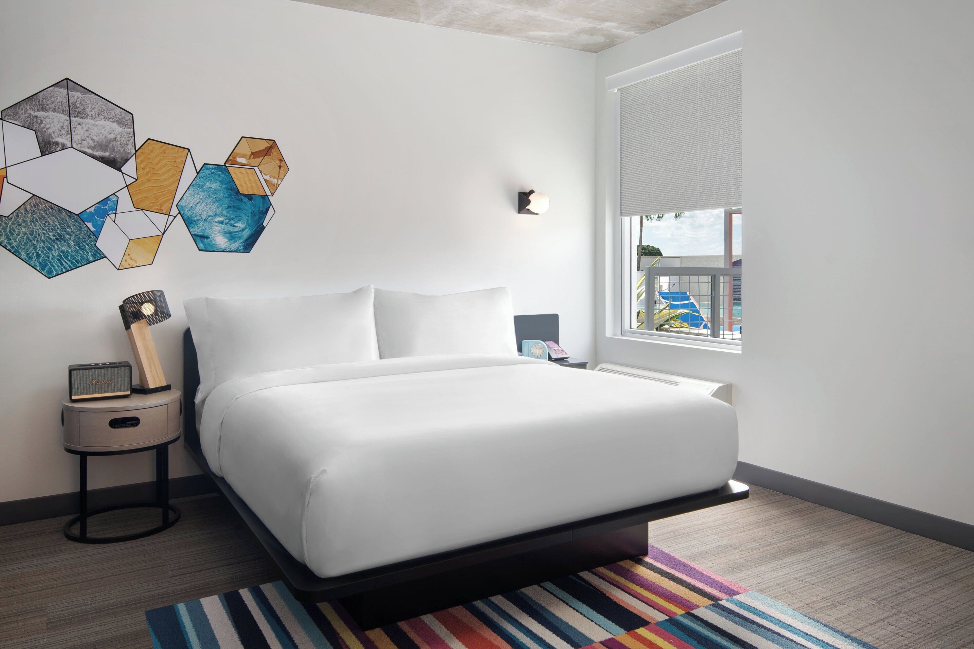 A modern hotel room with a large bed, a small bedside table with a radio, a colorful striped rug, and a geometric wall art piece.