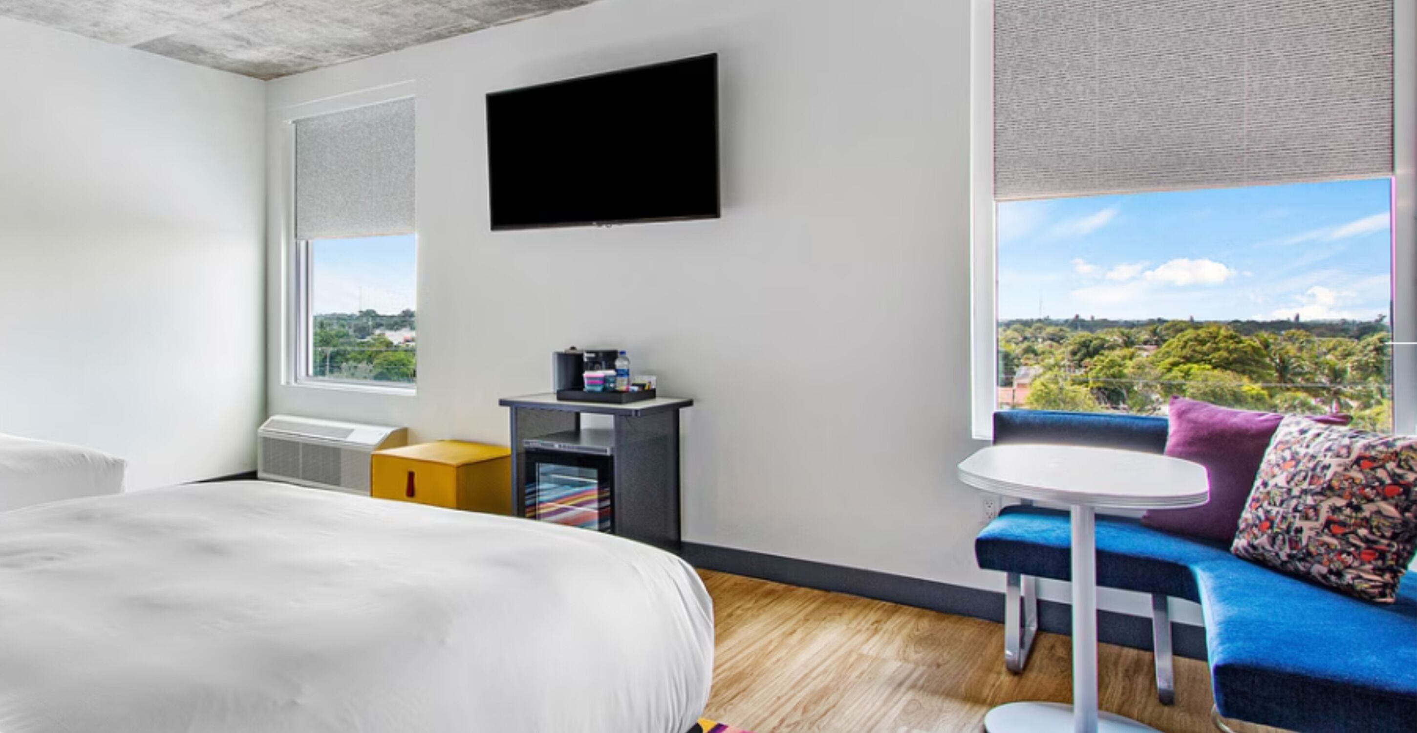 A hotel room with a bed, a TV mounted on the wall, a small table with a chair, and a sofa with cushions.