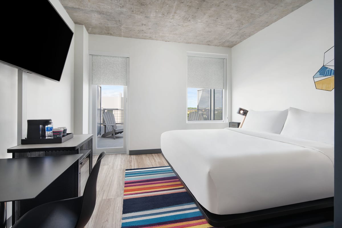 A modern hotel room with a large bed, a desk with a flat-screen TV, and a view of the city through the windows.